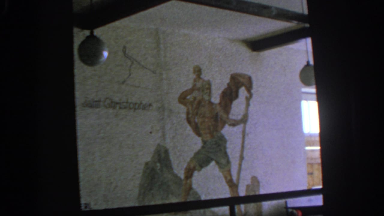 Textured wall with a Saint Christopher relief, inscription, hanging lamps, and dark film borders.