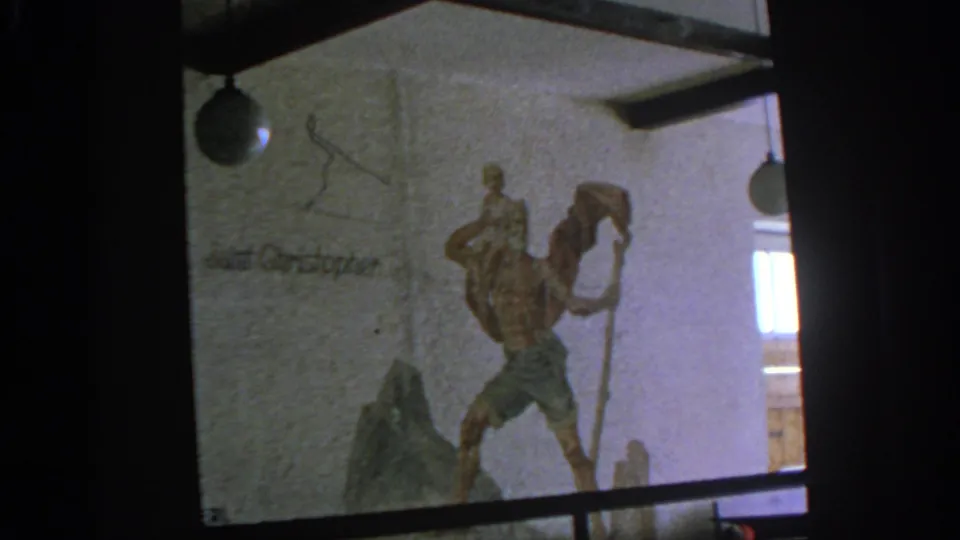 1963 8mm Munich-area home-movie frame showing a wall relief of Saint Christopher carrying a child, with lamps and partial inscription nearby.