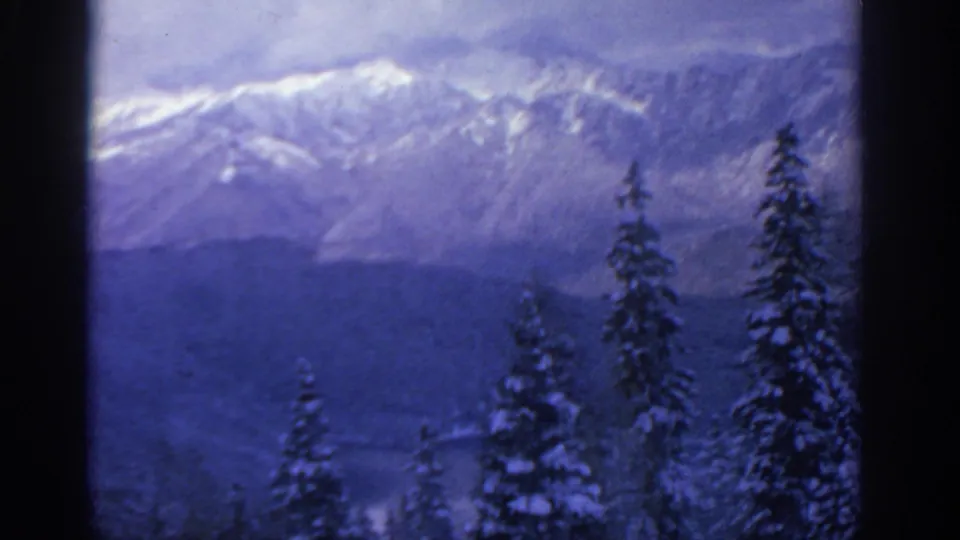 1963 8mm Munich-area scenic view of snow-dusted mountains beyond evergreen trees, rendered with soft grain and cool blue color.