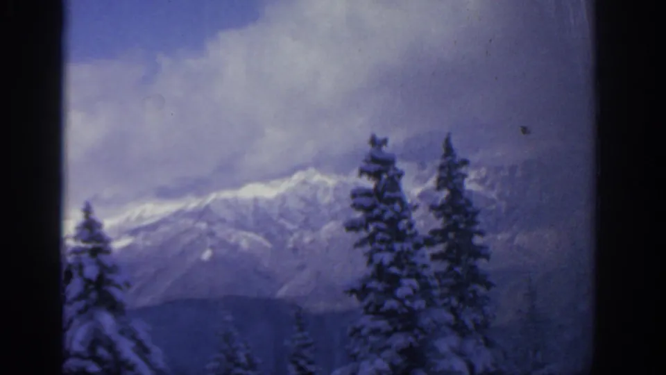 Munich-area 1963 8mm frame with dark film borders, snowy evergreens, and a clouded mountain backdrop.