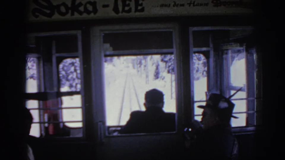 1963 home-movie frame inside a rail car, with passengers silhouetted against bright snow and trees beyond the windows.