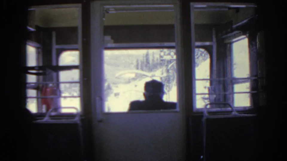 1963 Munich train interior with a passenger silhouette seated before bright snowy windows and carriage fixtures.