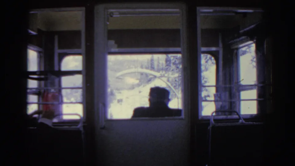 Inside a 1963 Munich train carriage, a dark passenger silhouette faces forward toward snowy trees and rail-side structures.