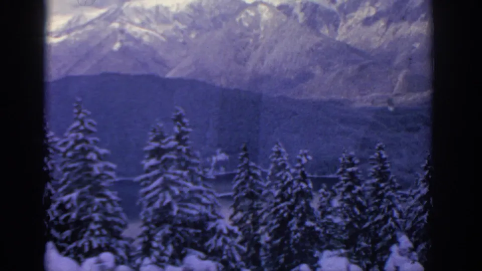 1963 Munich-area 8mm home-movie view of snowy evergreens and layered mountain ridges under a pale winter sky.
