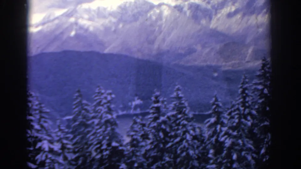 Soft 8mm home-movie frame from 1963 with snowy evergreens, dark foothills, and pale mountain ridges in winter light.