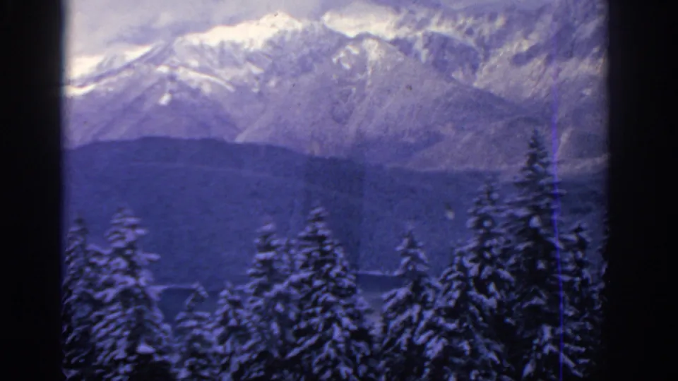 1963 8mm winter view near Munich with snow-covered treetops, dark wooded slopes, and distant snow-streaked alpine mountains.