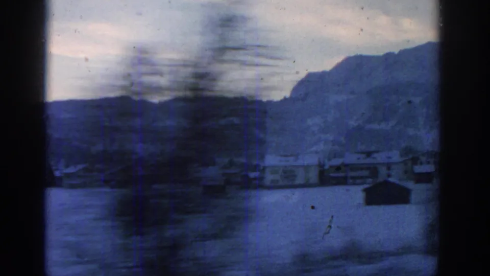 A passing 8mm travel view shows blurred foreground trees, small snow-covered buildings, and mountains in the distance.