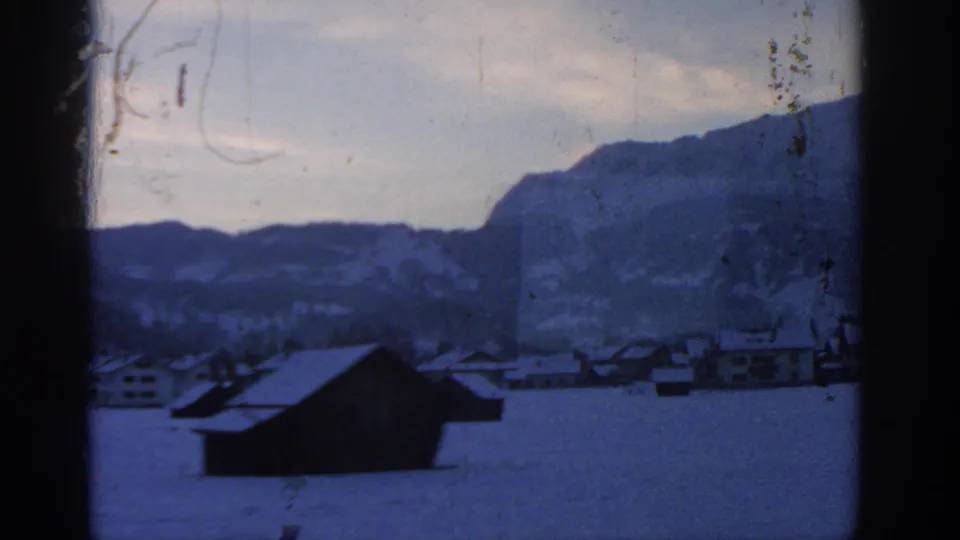 A dark barn-like structure stands in a snow-covered field, with low village buildings and mountain slopes in the background.