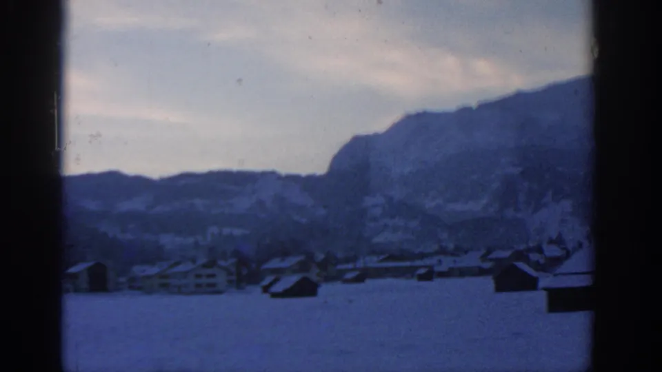 Snow-covered rooftops and small buildings lie below a blue-toned mountain ridge, with heavy window framing at the side.