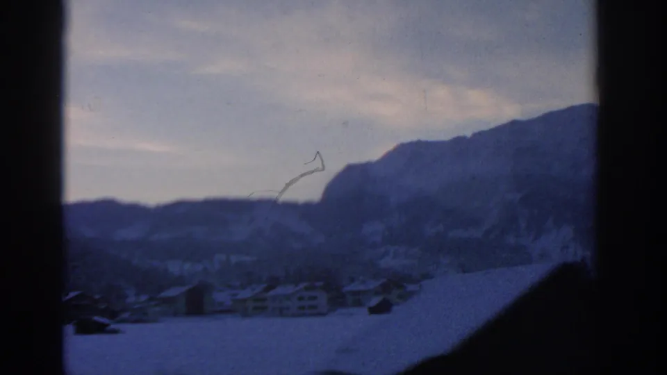 A soft blue 8mm frame shows a snowbound village below mountains, with dark foreground obstruction and visible film marks.