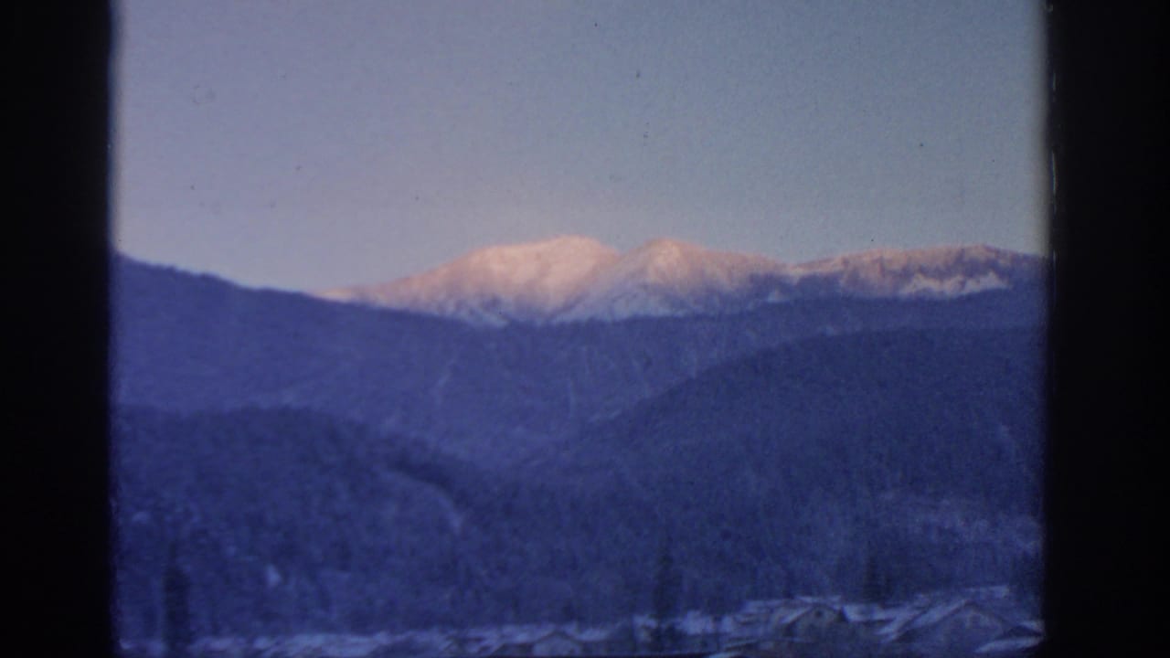 1963 Munich-area 8mm frame showing distant snow-covered peaks glowing softly above darker mountain ridges and valley rooftops.