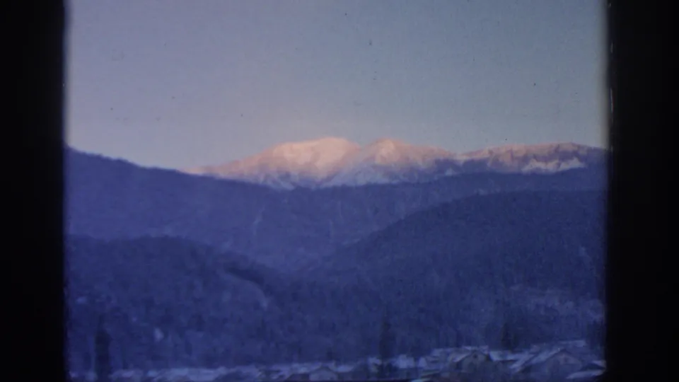 1963 Munich-area 8mm frame showing distant snow-covered peaks glowing softly above darker mountain ridges and valley rooftops.