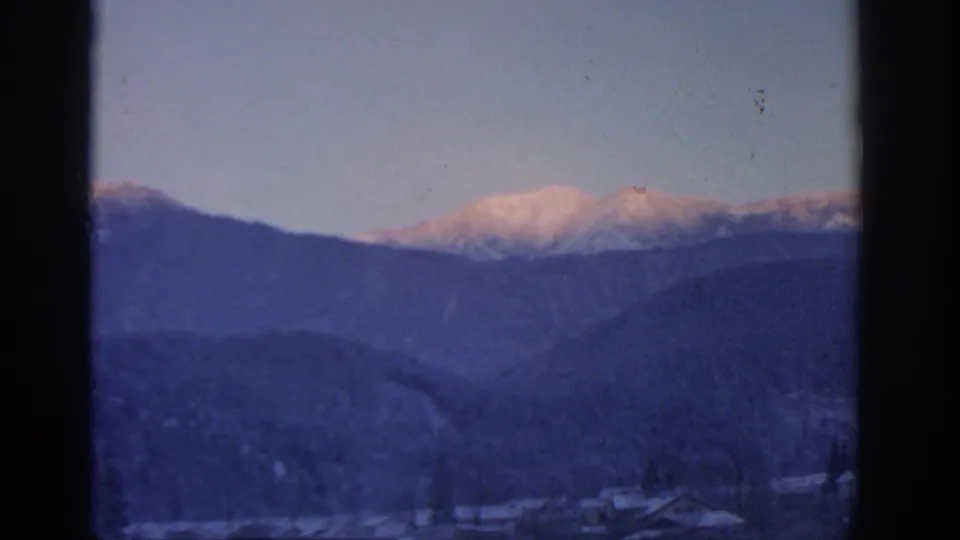 1963 Munich-area home-movie frame with a pink-lit snowy summit, layered blue ridges, pale sky, and dark 8mm frame borders.