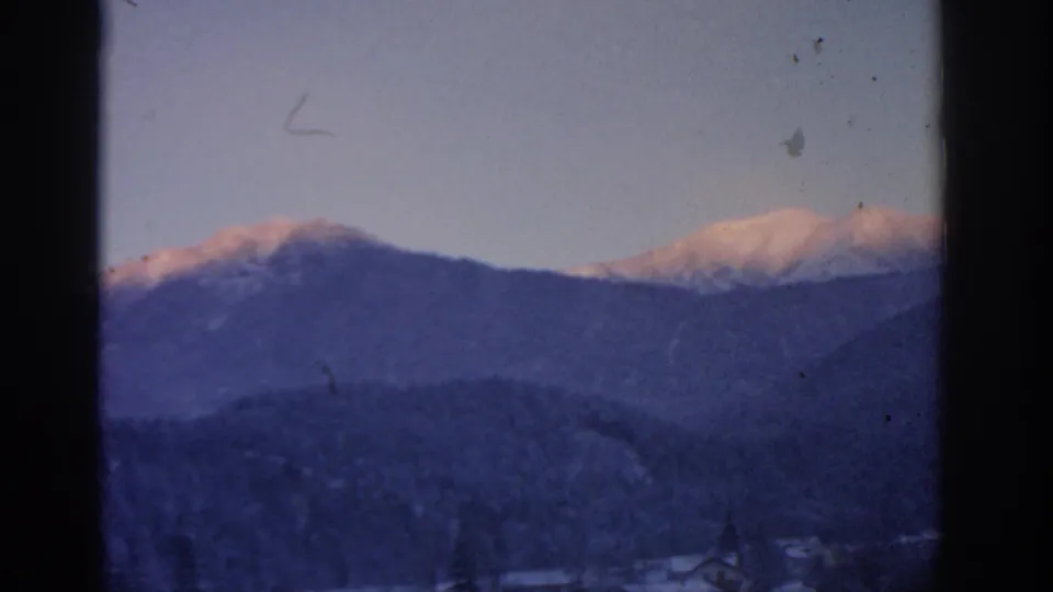 1963 Munich-area 8mm frame showing snow-bright mountain tops, dark wooded ridges, small valley buildings, and visible film dust.