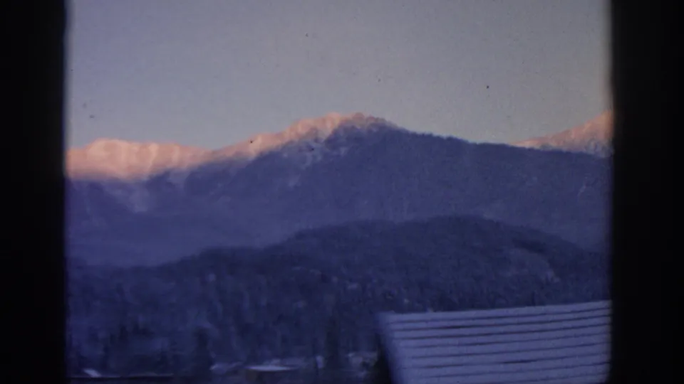 1963 Munich-area frame with layered winter mountains, pink snow highlights, pale sky, and a snow-covered roof at the lower right.