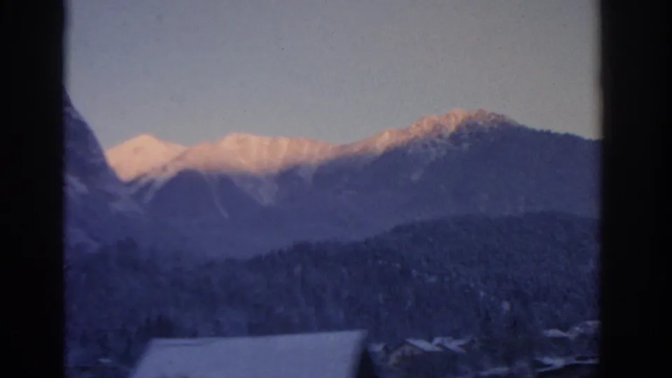 1963 8mm view near Munich with sunlit snowy peaks, dark vertical framing, and blurred winter village roofs below.