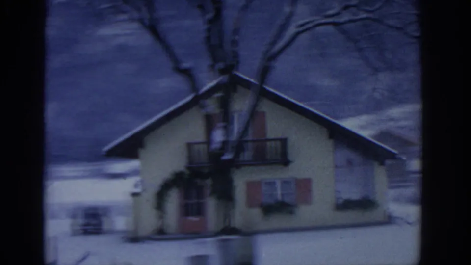 1963 8mm frame near Munich showing a snow-covered house, bare tree branches, a road edge, and a dark mountain backdrop.