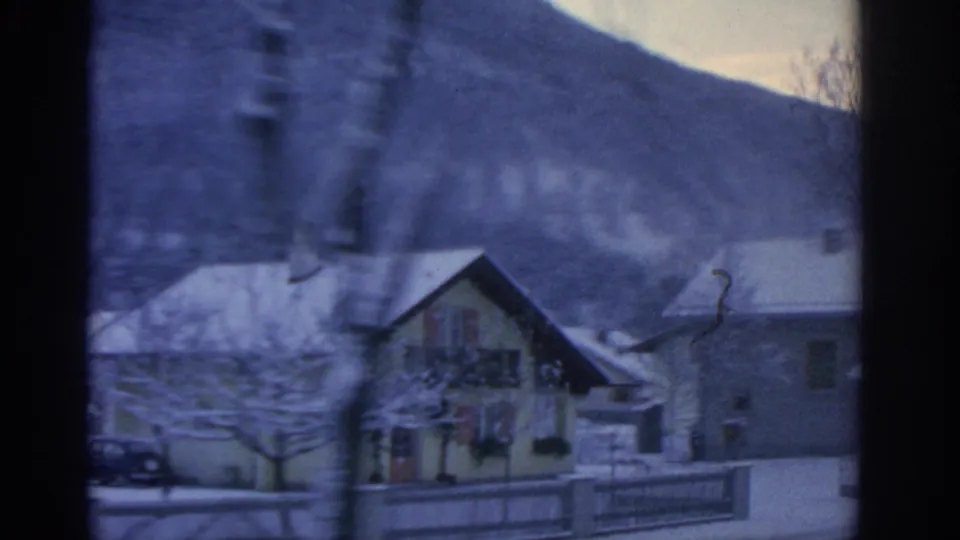 1963 8mm frame near Munich showing snowy village houses, a fence, bare winter trees, and a mountain slope beyond.