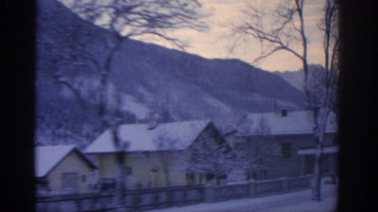 1963 8mm view near Munich, Germany, showing snowy houses, bare trees, and a fence from inside a moving vehicle.