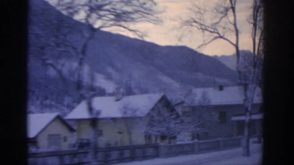 1963 8mm view near Munich, Germany, showing snowy houses, bare trees, and a fence from inside a moving vehicle.