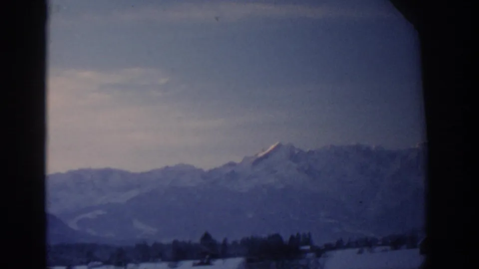 1963 8mm frame from the Munich travel clip showing distant snowy mountains and a dark foreground window edge.