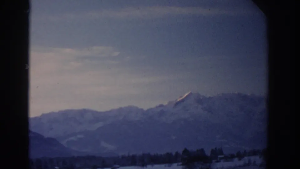 1963 8mm view near Munich with a snowy mountain range, low treeline, pale sky, and rounded home-movie frame edges.