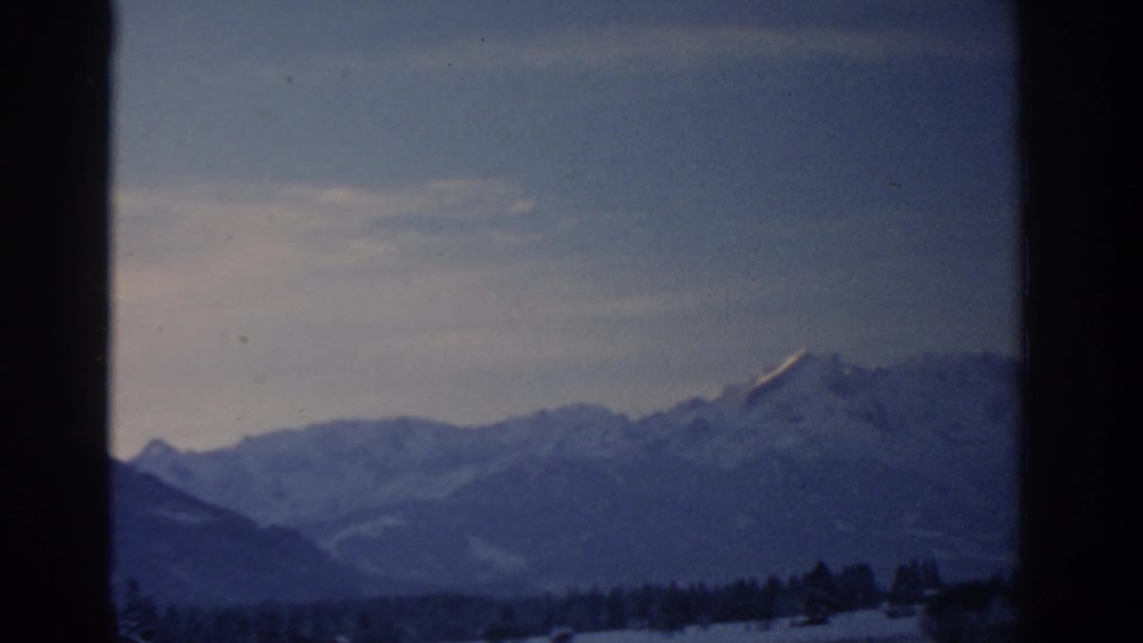 1963 8mm frame attributed to Munich, Germany, showing distant snowy mountains beneath a dim winter sky.