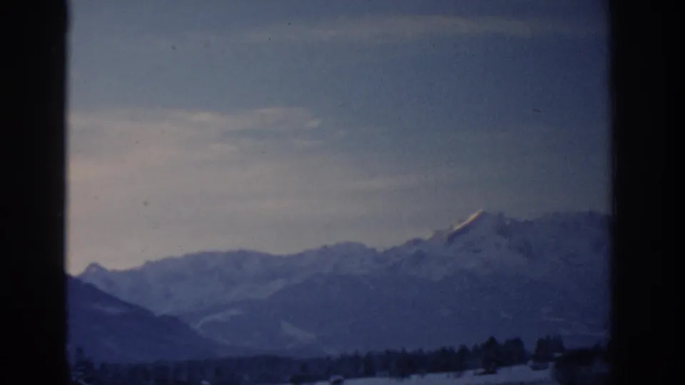 1963 8mm frame attributed to Munich, Germany, showing distant snowy mountains beneath a dim winter sky.
