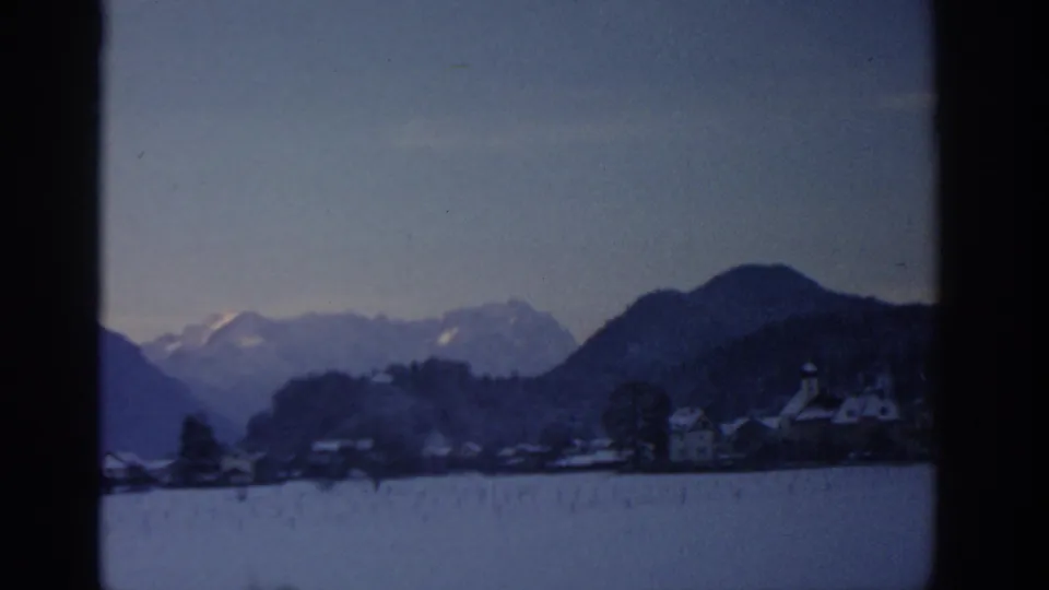 1963 8mm view attributed to Munich, Germany, showing a small snowy village below dark hills and distant mountains.