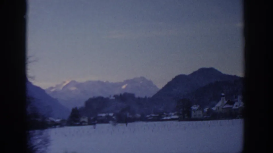 Late clip frame from 1963 8mm footage attributed to Munich, with a snowy village foreground and mountain ridges beyond.