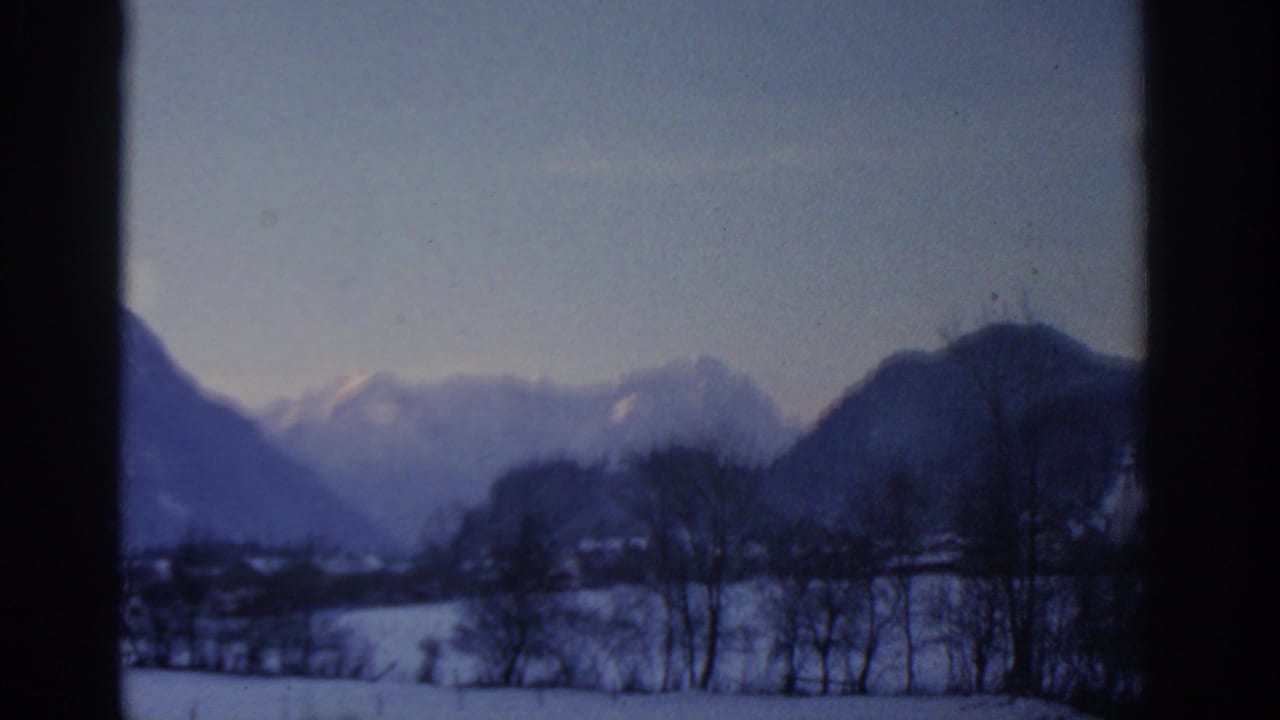 1963 8mm view near Munich showing snowy fields, bare trees and blue mountain silhouettes through a window.