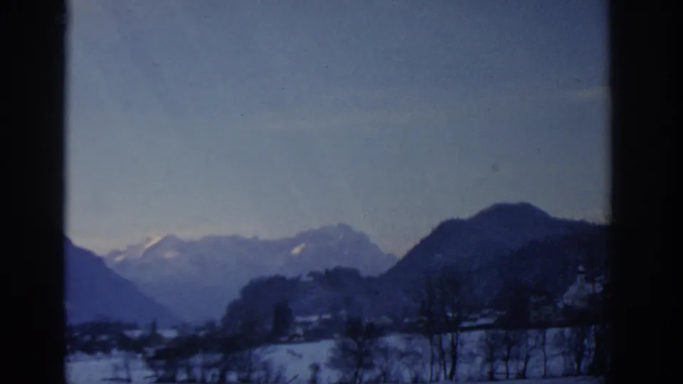 Window-framed 1963 Munich-area winter view with snowy fields, a small settlement and hazy mountain ridges.
