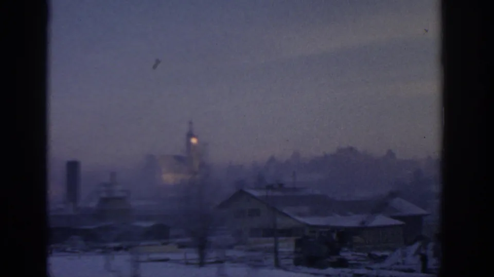 1963 Munich-area winter village view with a church steeple, rooftops, snow and hazy mountains beyond.
