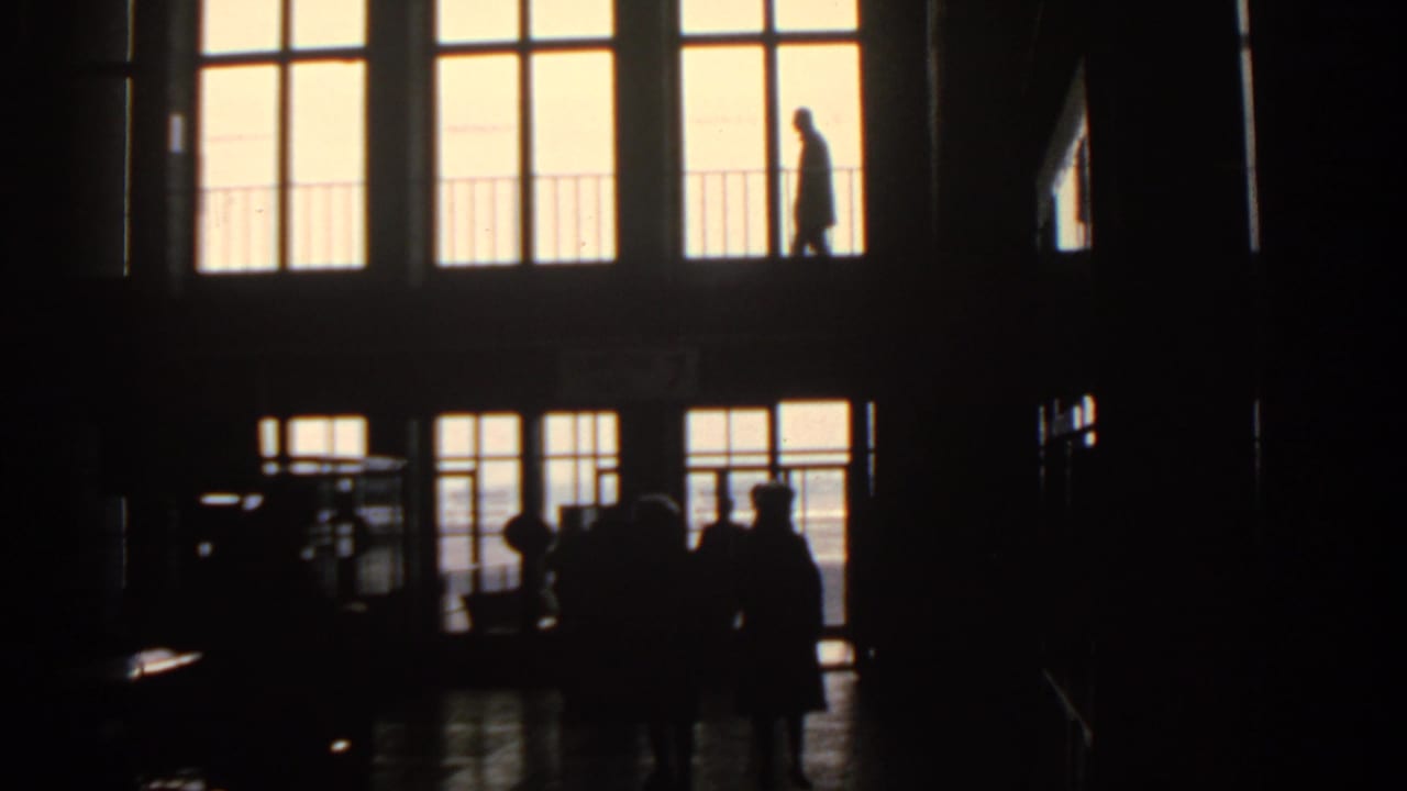 Silhouetted people stand and walk inside a dark concourse with bright glass doors.