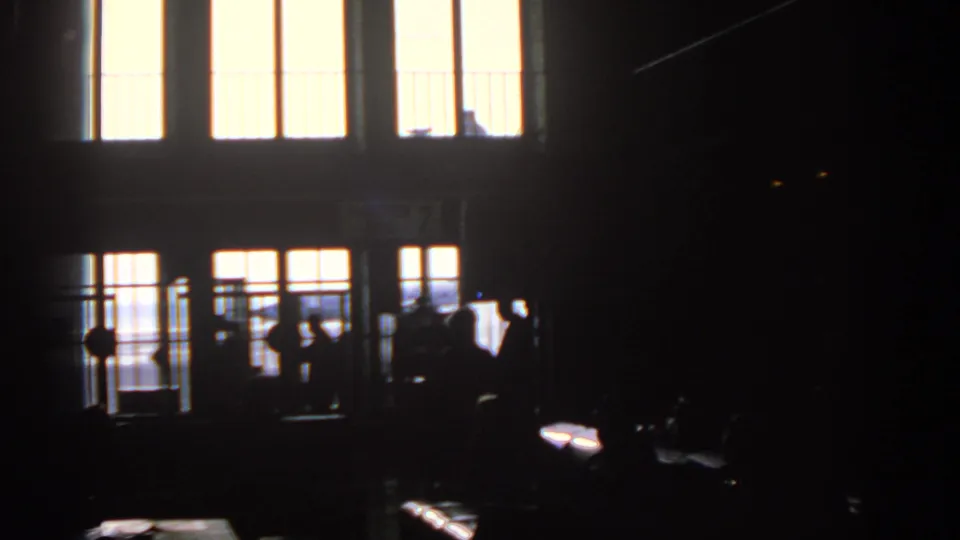A dim Munich airport terminal view shows silhouetted figures near gate railings and bright windows, with the 8mm exposure holding detail mostly in outline.