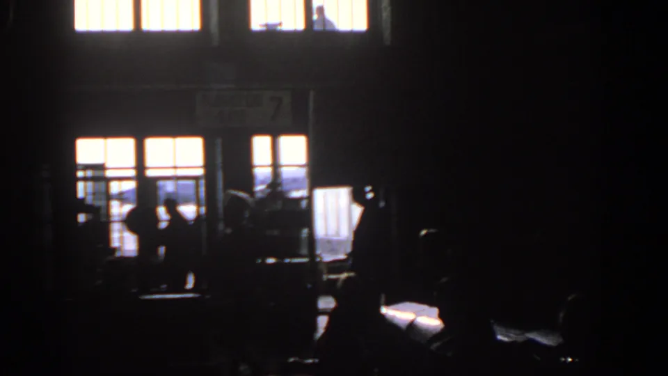 Travelers pass through a shadowed Munich airport gate area in 1963, with the Flugsteig 7 sign faintly visible above the entrance.