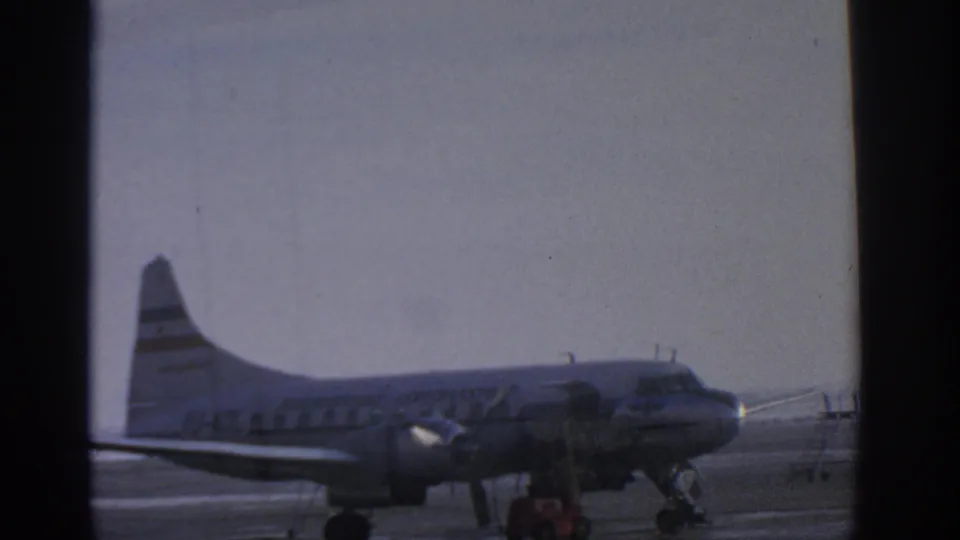 1963 Munich Airport view of a propeller passenger airplane with striped tail, seen through dark window mullions on the tarmac.