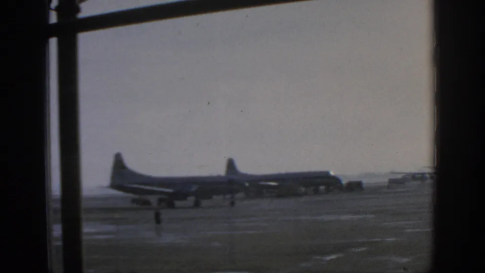 Wide 1963 Munich Airport view of distant propeller aircraft on the apron, with a small ground figure and window framing in the foreground.