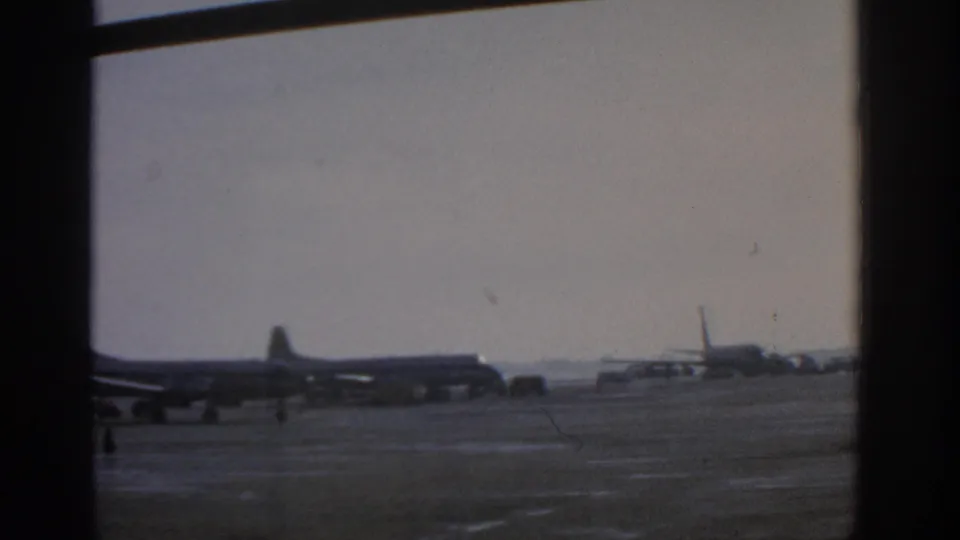 Wide home-movie view of Munich Airport in 1963, showing propeller aircraft, distant ground activity, and soft 8mm exposure through window framing.