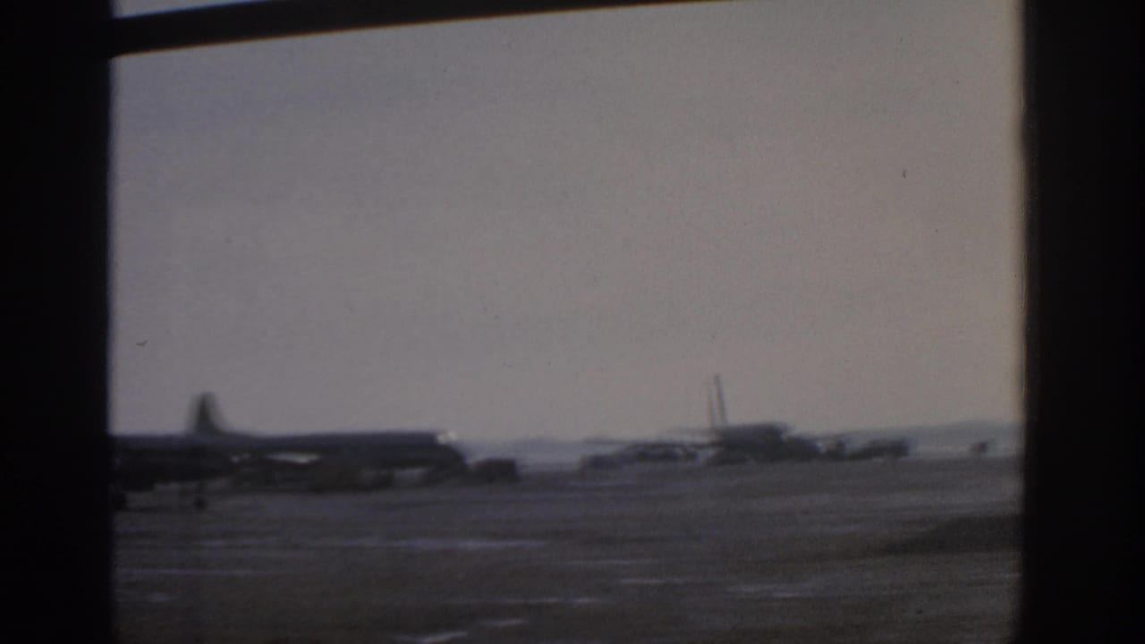 1963 Munich airport tarmac with propeller aircraft and service vehicles viewed through interior window mullions.