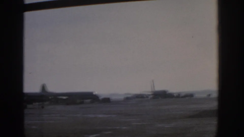1963 Munich airport tarmac with propeller aircraft and service vehicles viewed through interior window mullions.