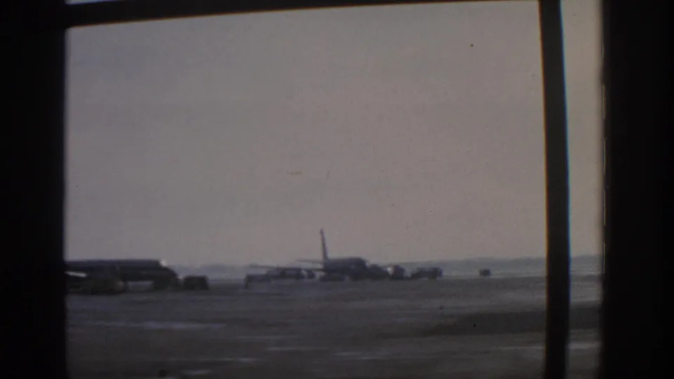 A Munich airport apron view shows parked commercial aircraft beyond heavy window framing in 1963 home-movie footage.
