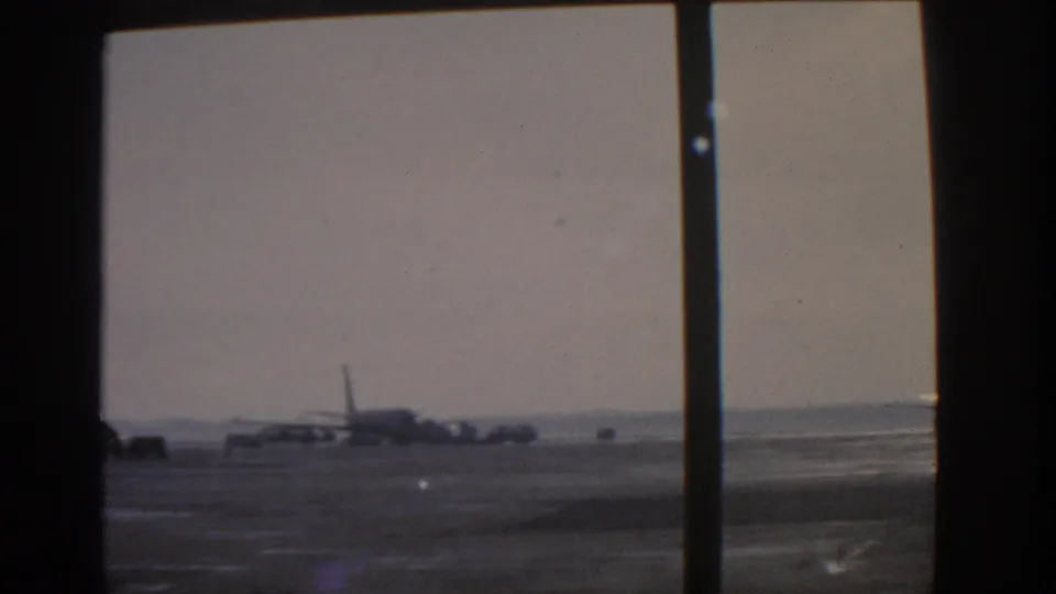 Wide 8mm view of the Munich airport tarmac in 1963, with aircraft and ground traffic partly obscured by window frames.