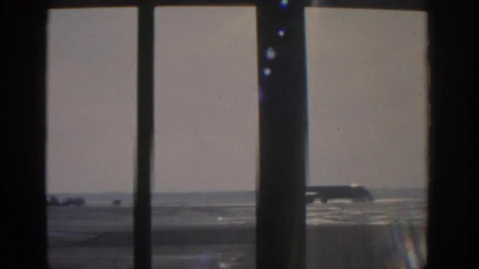 1963 Munich airfield view of a parked silver commercial airplane through dark window mullions on a bright day.