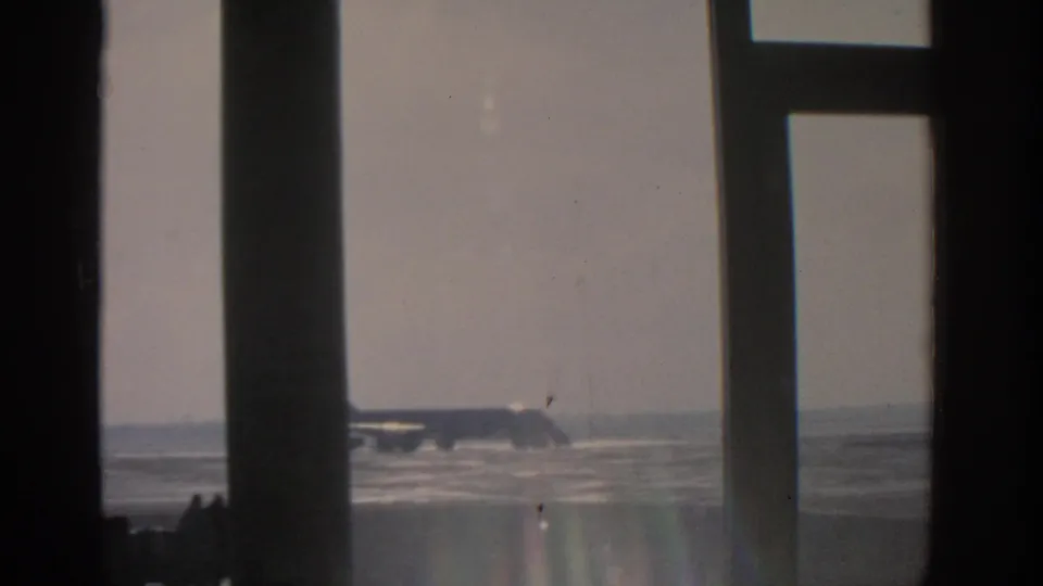 The parked silver aircraft appears partly hidden behind a dark window mullion in this 1963 Munich airport home-movie frame.
