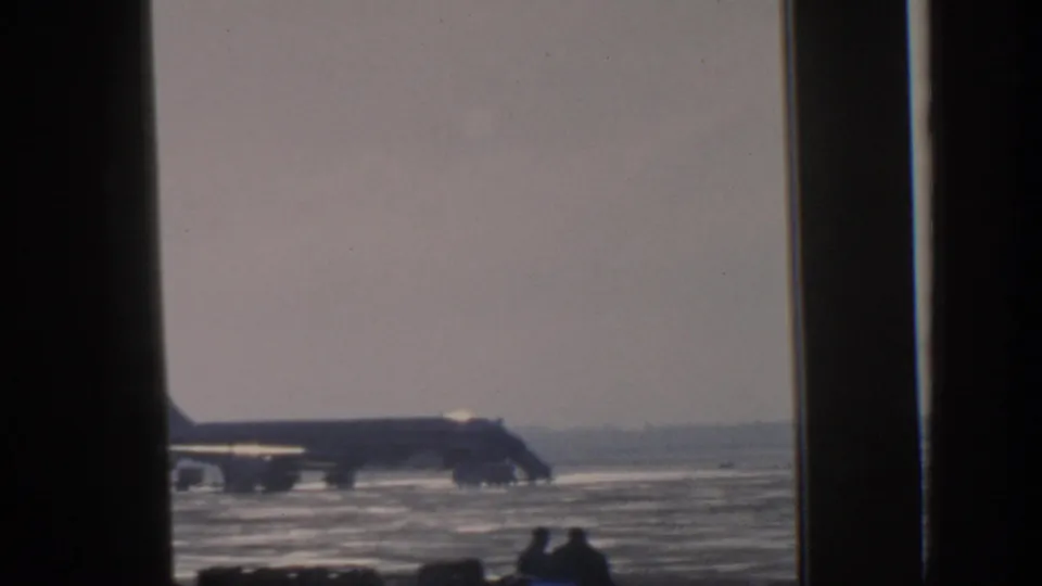 1963 Munich home-movie frame showing a distant airplane on a wet airfield through a dark interior window frame.