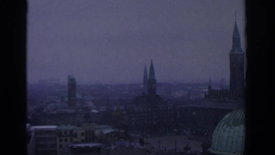Munich skyline in muted 1960s 8mm color, framed by dark vertical window bars and distant haze.