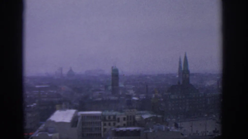 Munich city skyline in a brief 8mm home-movie shot, with distant towers, rooftops, and a pale hazy sky.