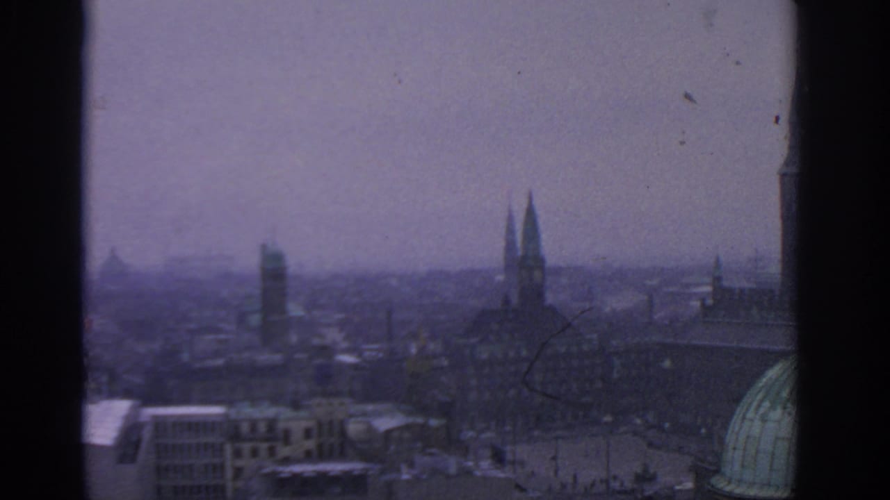 1963 8mm view over Warsaw, Poland, showing distant spires, dense rooftops, haze, and a green dome framed by dark borders.