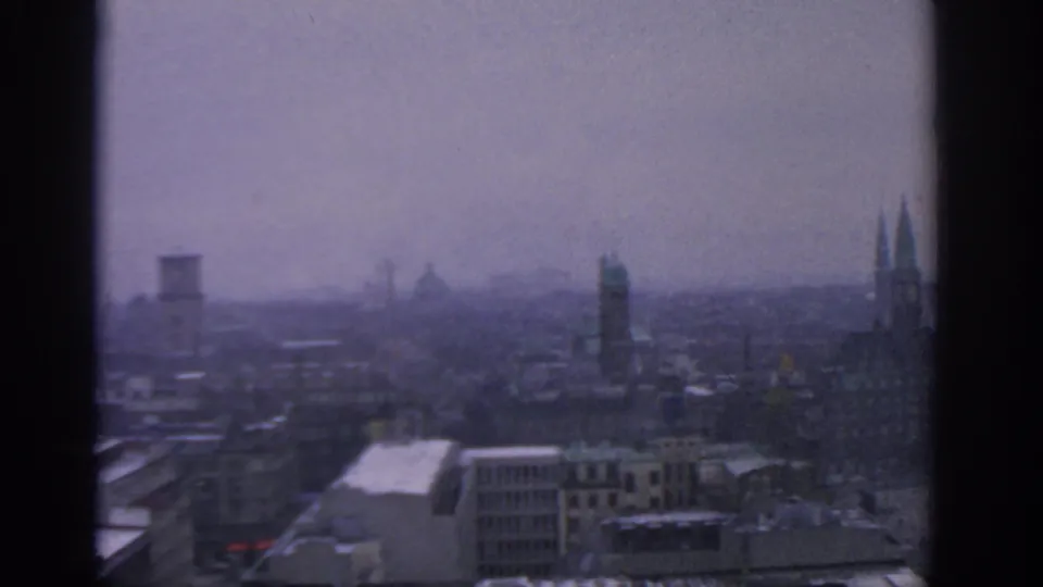 1963 Warsaw skyline seen from above, with distant towers, layered rooftops, soft haze, and dark 8mm frame borders.
